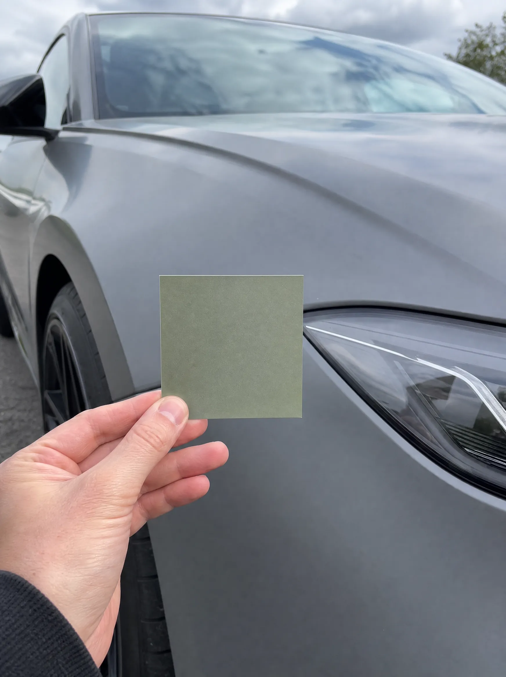 A hand holding a sage green film sample against a grey car wing
